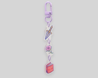 May include: A purple keychain with a lobster clasp, featuring charms of a dagger, a pink flower, and a book with the text "Enemies to Lovers". The charms are connected by small purple chains.