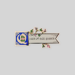 May include: A gold-toned metal enamel pin featuring a blue square with a stylized letter Q containing a pink and green diamond pattern. Attached is a cream-colored banner with the black text "queen of side quests" and decorative green vines.