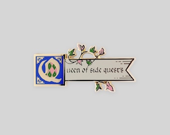 May include: A gold-toned metal enamel pin featuring a blue square with a stylized letter Q containing a pink and green diamond pattern. Attached is a cream-colored banner with the black text "queen of side quests" and decorative green vines.