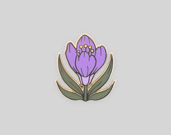 May include: A metal enamel pin shaped like a crocus flower. The flower features light purple petals with dark outlines, yellow stamens in the center, and dark green leaves at the base. The entire design is bordered by a thin gold-colored metal edge.