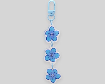 May include: A light blue keychain with three flower charms. Each charm is a blue flower with a white outline and a blue center. The keychain has a light blue clasp.