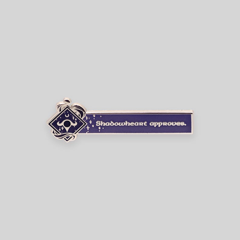May include: A silver enamel pin with a purple background and a silver design. The pin features a stylized symbol with a crescent moon and a star, and the text "Shadowheart approves."