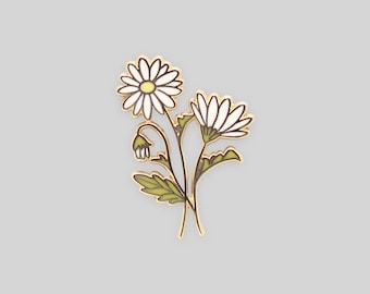 May include: A gold-toned metal enamel pin featuring two white daisy flowers with yellow centers, a small closed bud, and green leaves. The design is outlined in thin gold metal lines, creating a botanical illustration of a small flower bouquet.