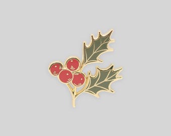 May include: A gold-toned metal enamel pin shaped like a sprig of holly. The design features three round, bright red berries clustered together next to two pointed, dark olive green leaves with gold outlines and detailed vein markings.