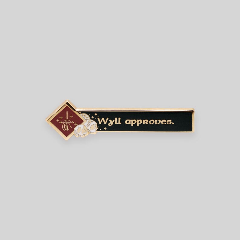 May include: Gold enamel pin with a red diamond shape featuring a stylized hand with a wand and the text "Wyll approves." The pin is on a white background.