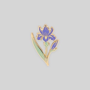 May include: A gold-toned metal enamel pin shaped like an iris flower. The design features a vibrant purple bloom with a pale yellow center, a light green stem and leaf, and a small purple bud, all outlined in polished gold metal lines.