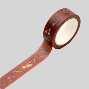 May include: A roll of decorative adhesive tape with a terracotta-colored background. The tape features a repeating pattern of cream-colored unicorns, small purple flowers, and green leafy vines that wind along the length of the tape surface.