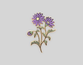 May include: A gold-toned metal enamel pin featuring a floral design with two blooming purple flowers, one small purple bud, and light green leaves. The petals are outlined in gold, and the center of each flower is a pale yellow circle.