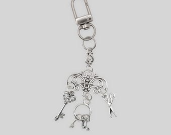 May include: Silver-colored keychain featuring a decorative filigree design with a key, scissors, and a small lock charm. The keychain has a lobster clasp for easy attachment. The design is intricate and detailed, with a vintage aesthetic.