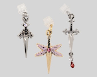 May include: Three metal charms with clear plastic earring backs. The left charm is a silver dagger with a crescent moon and iridescent stone. The center is a gold dragonfly-shaped dagger with pink wings and a pink stone. The right is a silver sword.