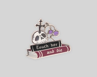 May include: A metal enamel pin featuring a stack of two books with a skull pierced by a sword and a sprig of purple flowers. The top book spine is black with the white text Touch her, and the bottom book is maroon with the white text and die.