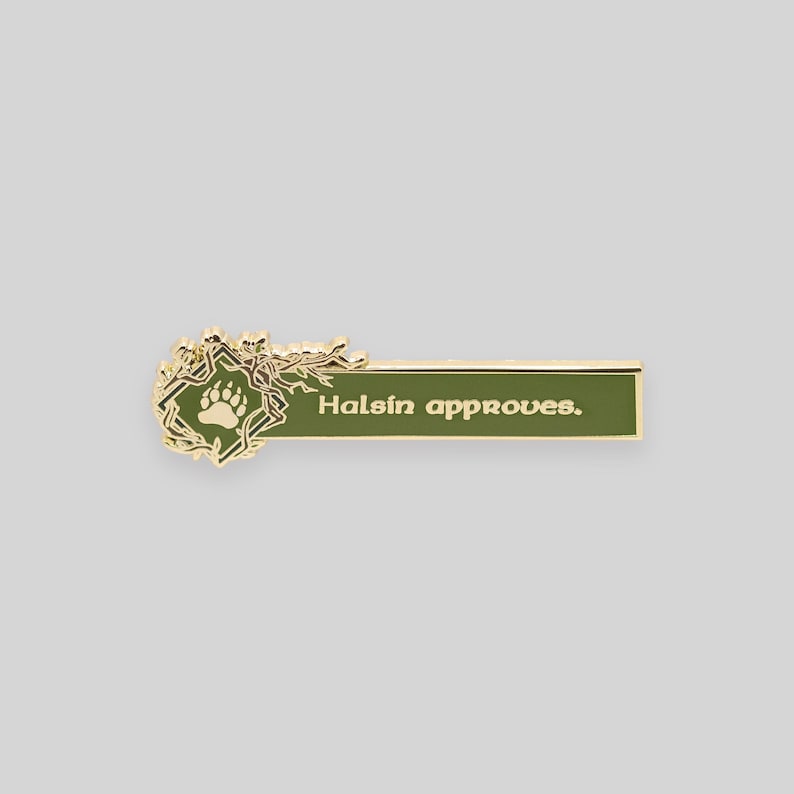 May include: Gold-colored enamel pin with a green background. The pin features a stylized tree branch with a bear paw print and the text "Halsin approves."