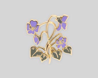 May include: A gold-toned metal enamel pin featuring a cluster of purple violet flowers with yellow centers and dark green heart-shaped leaves. The design showcases delicate stems and multiple blossoms arranged in a botanical floral pattern.