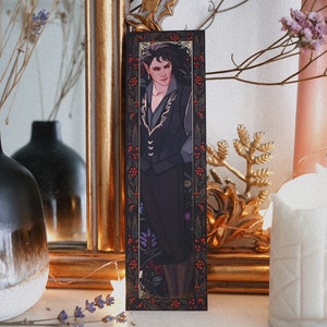 May include: A black bookmark with a gold border and a floral design. The bookmark features a portrait of a person with dark hair and a black jacket. The person is wearing a gold necklace and has a serious expression.