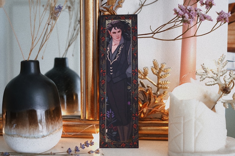 May include: A black bookmark with a gold border and a floral design. The bookmark features a portrait of a person with dark hair and a black jacket. The person is wearing a gold necklace and has a serious expression.