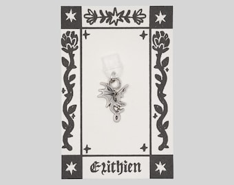 May include: A silver-toned metal dragon charm attached to a small clear plastic backing. The charm is displayed on a rectangular white card featuring a black ink border with floral motifs and the text Elithien printed at the bottom center.