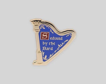 May include: A gold-toned metal enamel pin shaped like a harp. The harp features a blue background with white decorative floral accents and a red square containing a white letter S. The text reads Seduced by the Bard in white lettering.