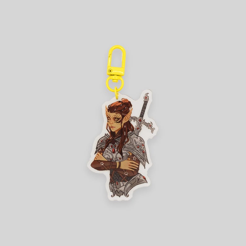 May include: A clear acrylic keychain featuring a detailed illustration of a female warrior with brown hair, wearing armor and holding a sword. The keychain has a yellow metal clasp.