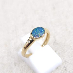 May include: A gold ring featuring an oval opal with blue and green hues. A small diamond accent is set on the band. The ring is displayed on a white stand against a neutral background.