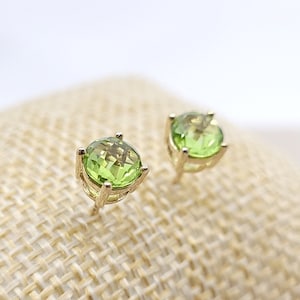 May include: A pair of gold earrings with green gemstones. The earrings are on a beige fabric background.