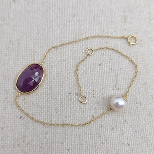 May include: A fine gold bracelet with a large, oval-cut ruby and a white pearl. The ruby is set in a gold bezel, and the pearl is round. The bracelet has a delicate chain and a clasp.