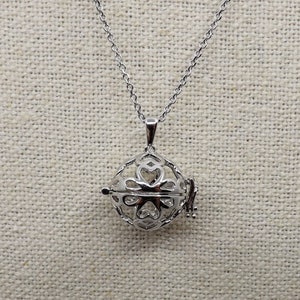 Op de afbeelding: Zilverkleurige hangerketting met een ronde, opengewerkte kooi-ontwerp met hartvormige uitsparingen. De kooi opent om een witte binnenkant te onthullen. De ketting heeft een fijne ketting en een eenvoudige sluiting.