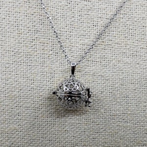 以下が含まれることがあります： 繊細な透かし彫りの球形のペンダントが付いたシルバーネックレス。ペンダントには小さなヒンジ付きの開口部があります。