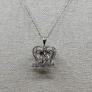 Könnte beinhalten: Eine silberne Halskette mit einem zarten Kronenanhänger, der mit einem filigranen Design und kleinen klaren Steinen verziert ist.