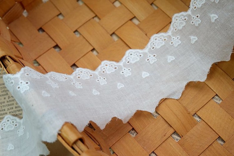 Peut inclure: Bordure en dentelle de coton blanche avec un bord festonn&eacute; et de petites fleurs brod&eacute;es. La dentelle est pr&eacute;sent&eacute;e sur un panier tiss&eacute;.