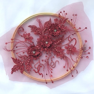May include: A burgundy floral applique with sequins and beads, arranged on a wooden embroidery hoop and pink mesh fabric. The design features detailed flower and leaf patterns, with sparkling embellishments.