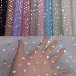 Puede incluir: Una variedad de telas de tul coloridas con lunares iridiscentes. Las telas están enrolladas, mostrando tonos de negro, marrón, lavanda, rosa, azul y blanco. Una mano sostiene un trozo de tul blanco con grandes puntos brillantes.