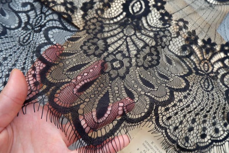 May include: Black lace fabric with a delicate floral pattern. The lace has a scalloped edge and is sheer.