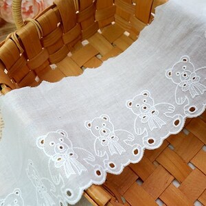 Peut inclure: Bordure en dentelle de coton blanche avec un bord festonn&eacute; et un motif r&eacute;p&eacute;t&eacute; d'ours en peluche brod&eacute;s. La dentelle est pr&eacute;sent&eacute;e dans un panier tiss&eacute;.