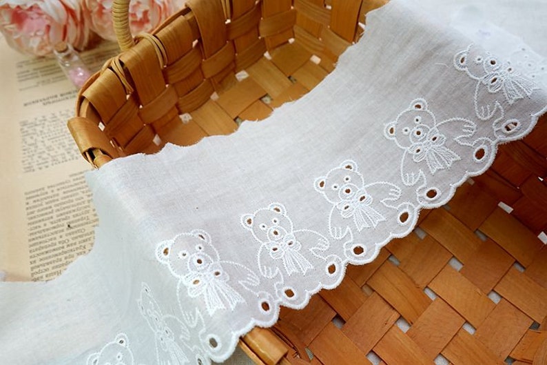 Peut inclure: Bordure en dentelle de coton blanche avec un bord festonn&eacute; et un motif r&eacute;p&eacute;t&eacute; d'ours en peluche brod&eacute;s. La dentelle est pr&eacute;sent&eacute;e dans un panier tiss&eacute;.