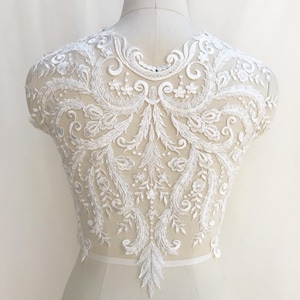 Può includere: Applique in pizzo bianco con un motivo floreale. Il pizzo è delicato e intricato, con una varietà di motivi floreali. L'applique è perfetta per aggiungere un tocco di eleganza a qualsiasi capo.