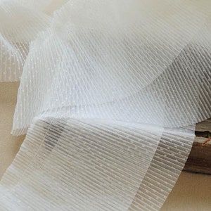 May include: White, sheer, pleated fabric with a dotted texture. The fabric is folded and layered, showing the texture and pleats.
