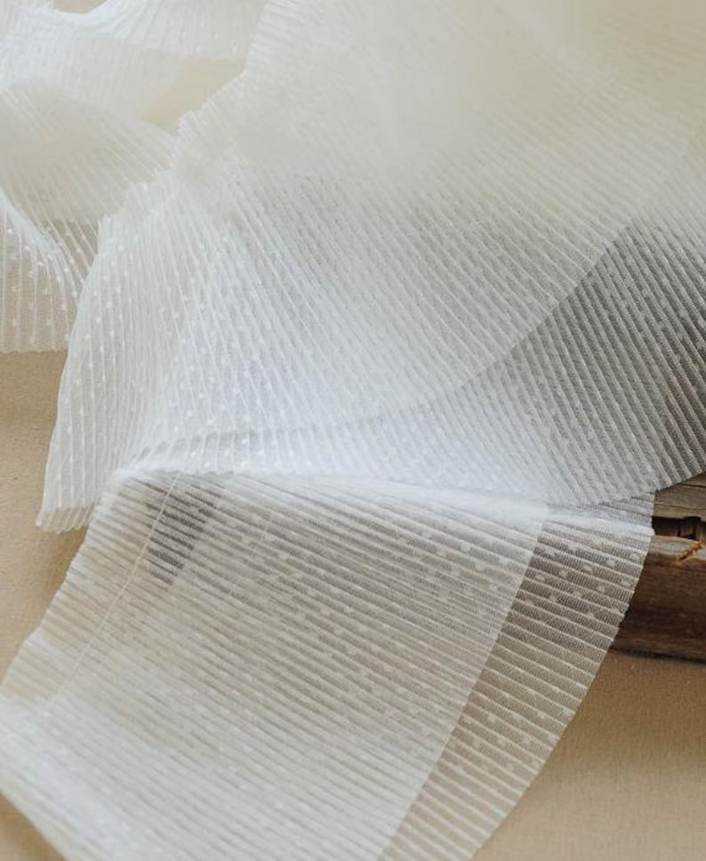 May include: White, sheer, pleated fabric with a dotted texture. The fabric is folded and layered, showing the texture and pleats.