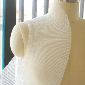 May include: White sheer fabric with a dotted pattern and pleated detail. The fabric is draped over a dress form.