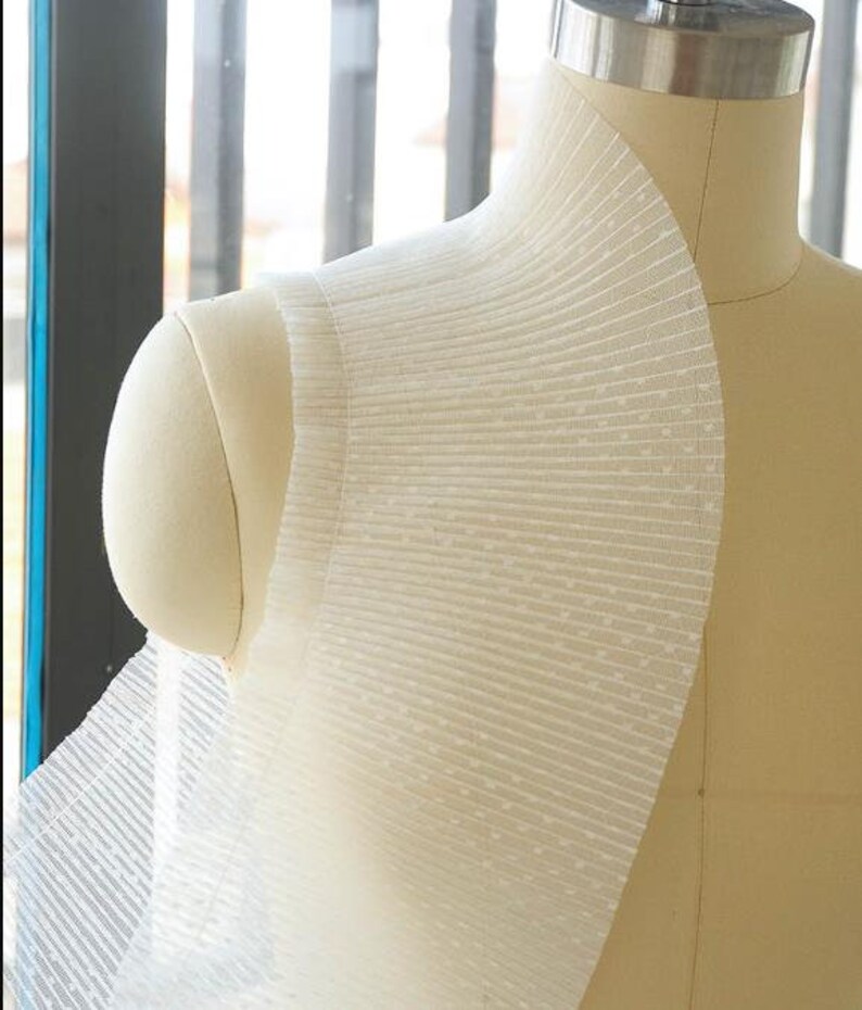 May include: White sheer fabric with a dotted pattern and pleated detail. The fabric is draped over a dress form.