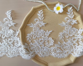 May include: White lace trim with a delicate floral pattern. The lace is shown on a beige surface with a white pearl necklace and a white flower.