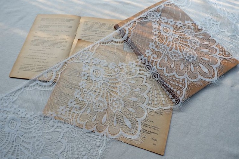 May include: White lace trim with a delicate floral pattern. The lace is draped over a book with a brown cover.