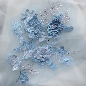 Könnte beinhalten: Hellblaue Blumenapplikation mit Pailletten und Perlen. Die Applikation zeigt eine Ansammlung von Blumen mit zarten Blättern und Stielen. Die Applikation ist perfekt, um jedem Projekt einen Hauch von Eleganz zu verleihen.