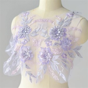 May include: A delicate lavender floral applique with pearl accents on a sheer mesh fabric. The applique features 3D flowers and leaves, perfect for adding a touch of elegance to clothing or crafts.