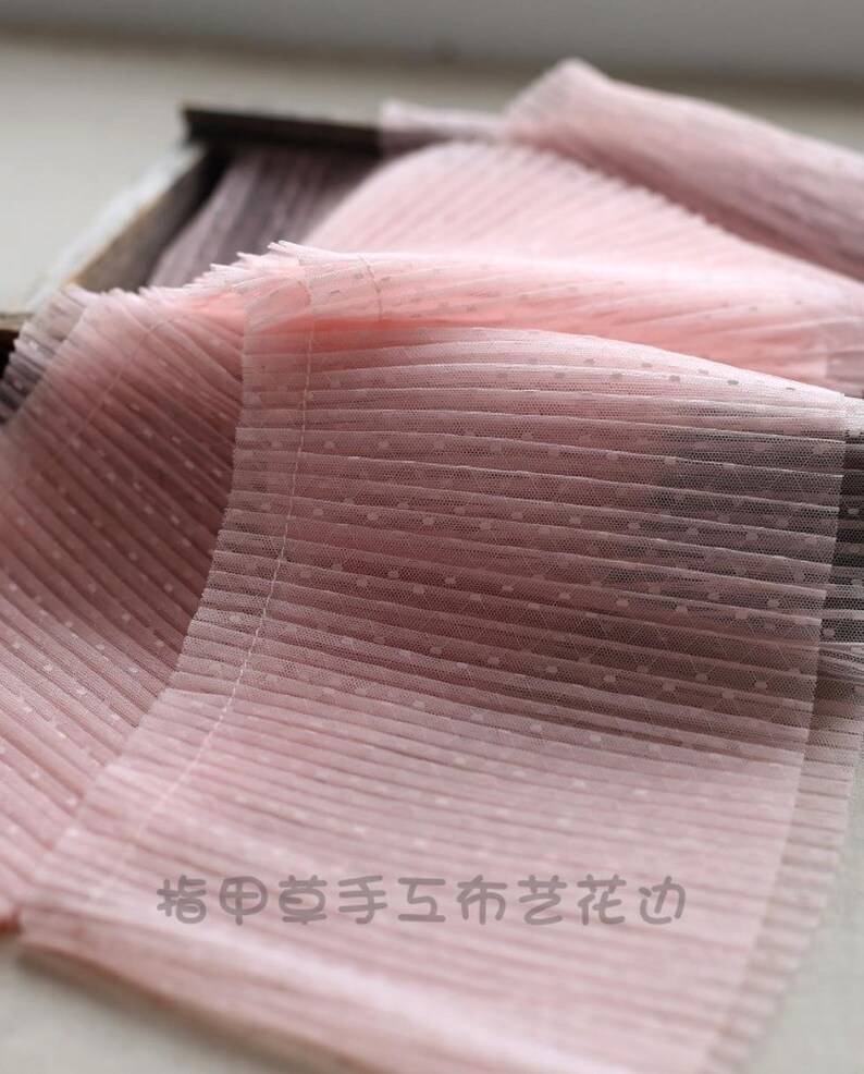 May include: Pink pleated tulle fabric with small white polka dots. The fabric is folded and has a soft, delicate texture.