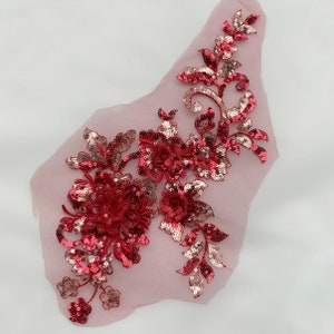 Peut inclure: Applique florale rouge et or avec des paillettes sur un tissu de maille transparent. L'applique présente une grande fleur avec des fleurs plus petites et des feuilles.
