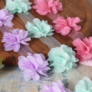 May include: A close-up of a ribbon with pastel-colored fabric flowers. The ribbon is made of a sheer, delicate material and the flowers are arranged in a repeating pattern. The flowers are in shades of pink, purple, and green.