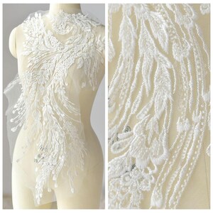 May include: White lace applique with a delicate floral design. The applique features a large, intricate pattern of feathers and leaves. It is perfect for adding a touch of elegance to any garment.