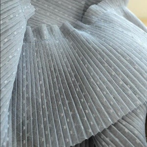 May include: Close-up of a light blue pleated fabric with white polka dots. The fabric is soft and delicate, and the pleats are evenly spaced.