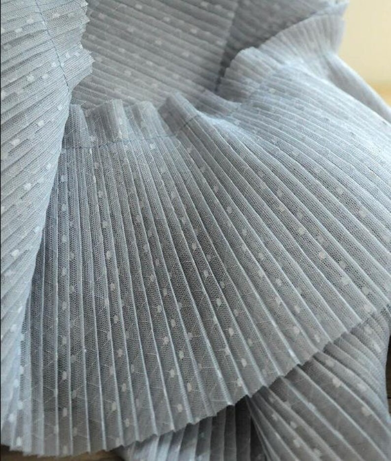 May include: Close-up of a light blue pleated fabric with white polka dots. The fabric is soft and delicate, and the pleats are evenly spaced.