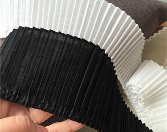 May include: Two rolls of pleated fabric trim, one black and one white. The fabric is gathered into pleats and has a stitched edge.